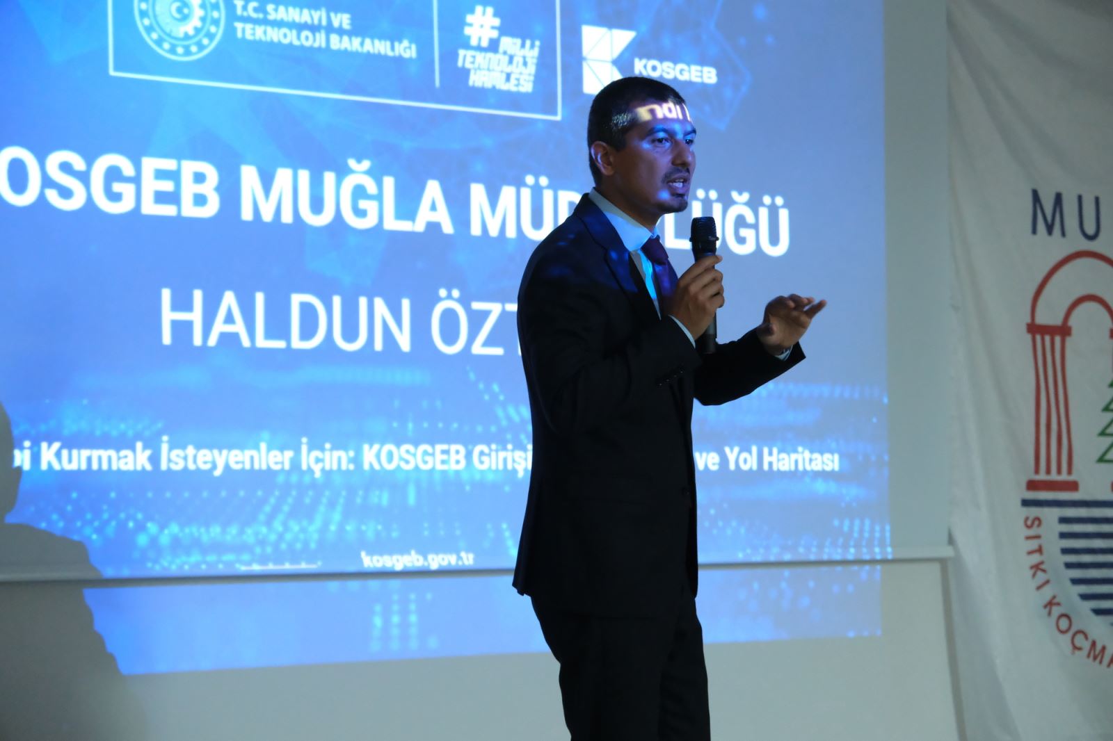  During the conference, the speaker is delivering a presentation to participants via a microphone in front of a projection screen.